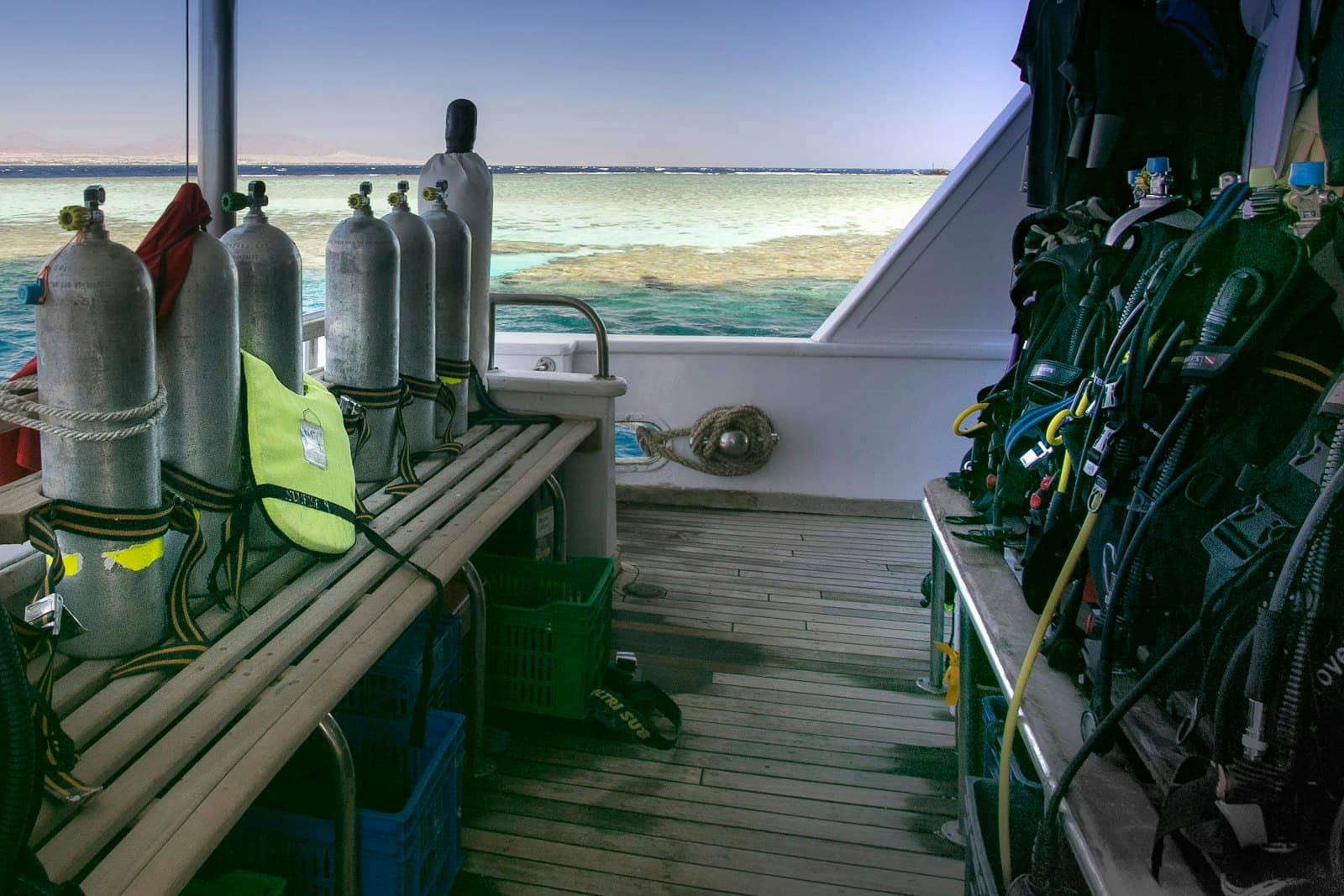 The dive deck of a Red Sea day boat: tanks strapped along the bench, BCDs hung and ready, the reef just off the stern.